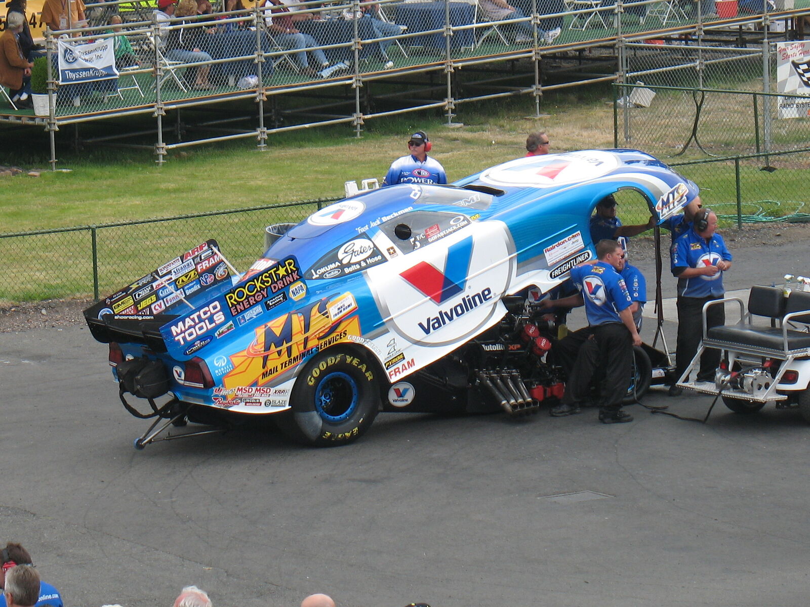 Jack Beckman's blue and white Valvoline / Mail Terminal Services Dodge Charger Funny Car with its body raised, crew members in blue Valvoline shirts working at the front of the car, grandstand fans behind a fence, Pacific Raceways, July 18, 2008